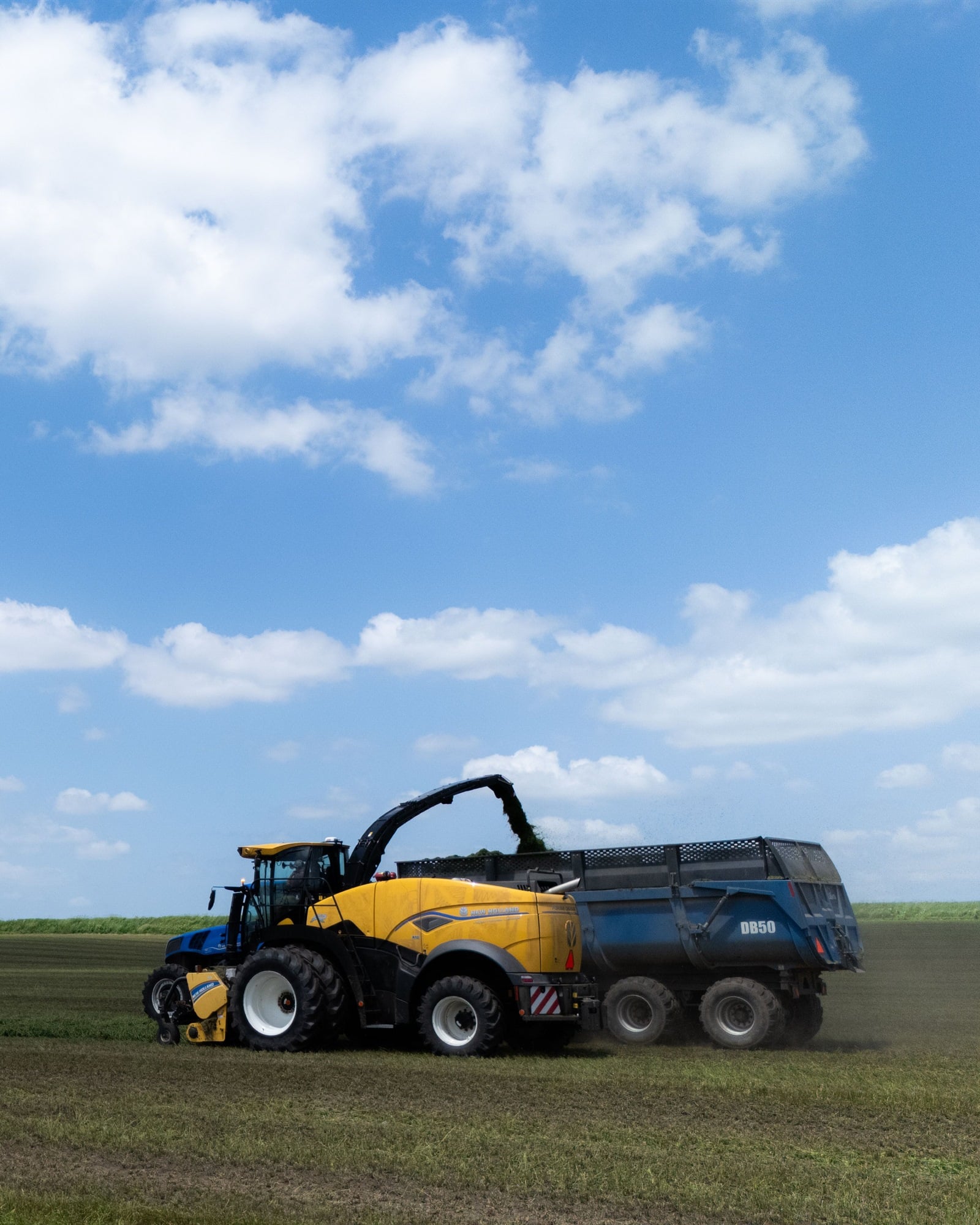 A forage harvester blowing silage into a trailer in a mown field under a summer sky